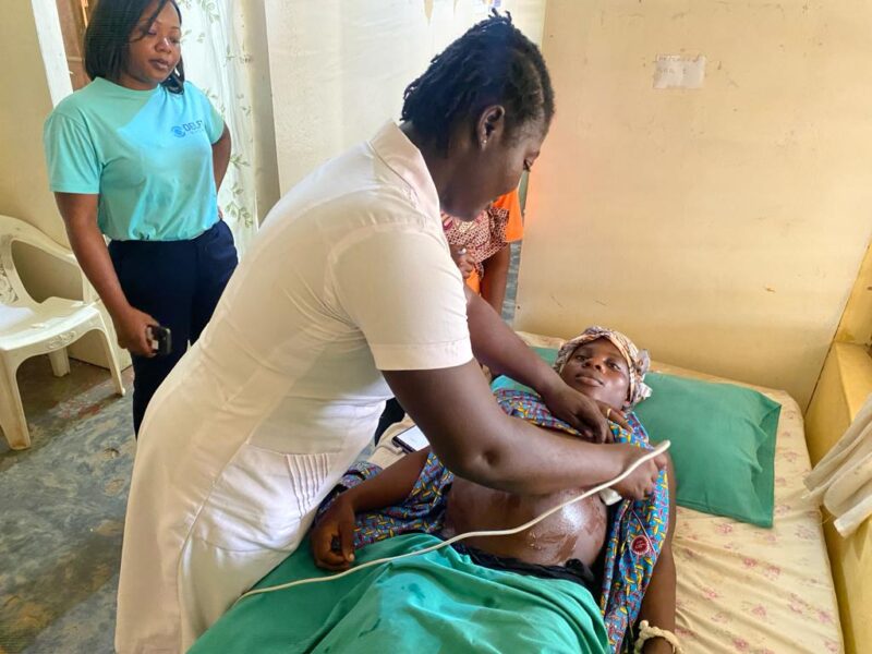 A health worker performing a BabyChecker obstetric ultrasound scan on a pregnant patient at a health facility in Ghana, observed by a Delft Imaging representative.
