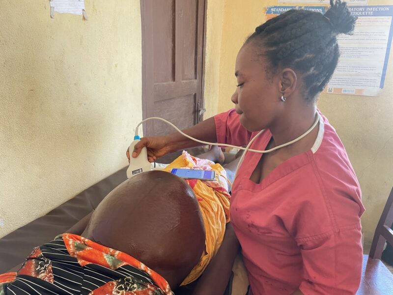 A health worker using BabyChecker to perform an obstetric ultrasound scan on a pregnant woman at a health facility in Sierra Leone, with the BabyChecker app visible on a smartphone.